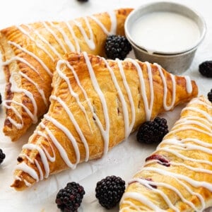 Blackberry Cream Cheese Turnovers on a Pan with Glaze and a Few Blackberries on a White Counter.