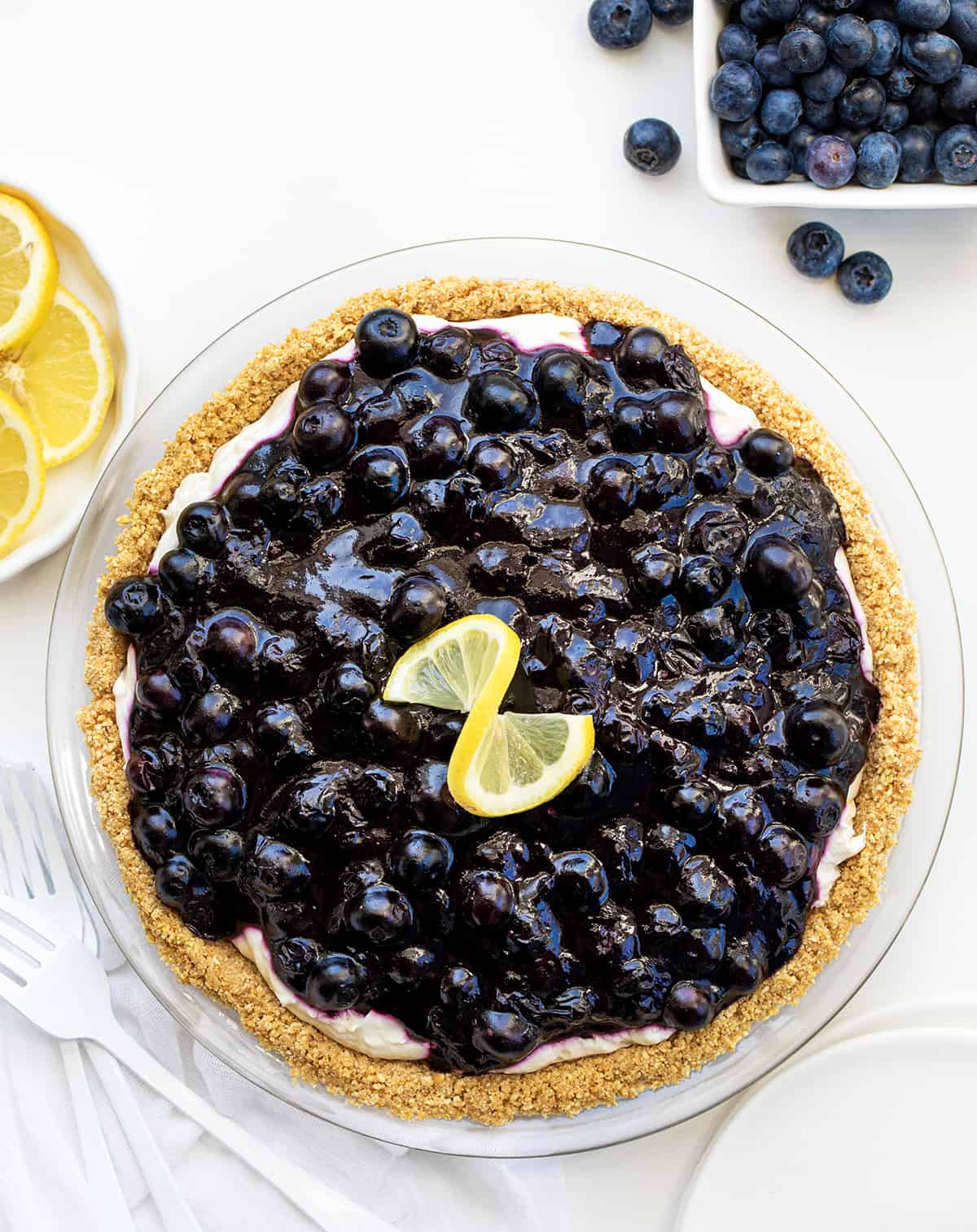 Looking Down on a Blueberry Lemon Cream Pie on a White Counter Surrounded by Blueberries and Lemons and White Forks.