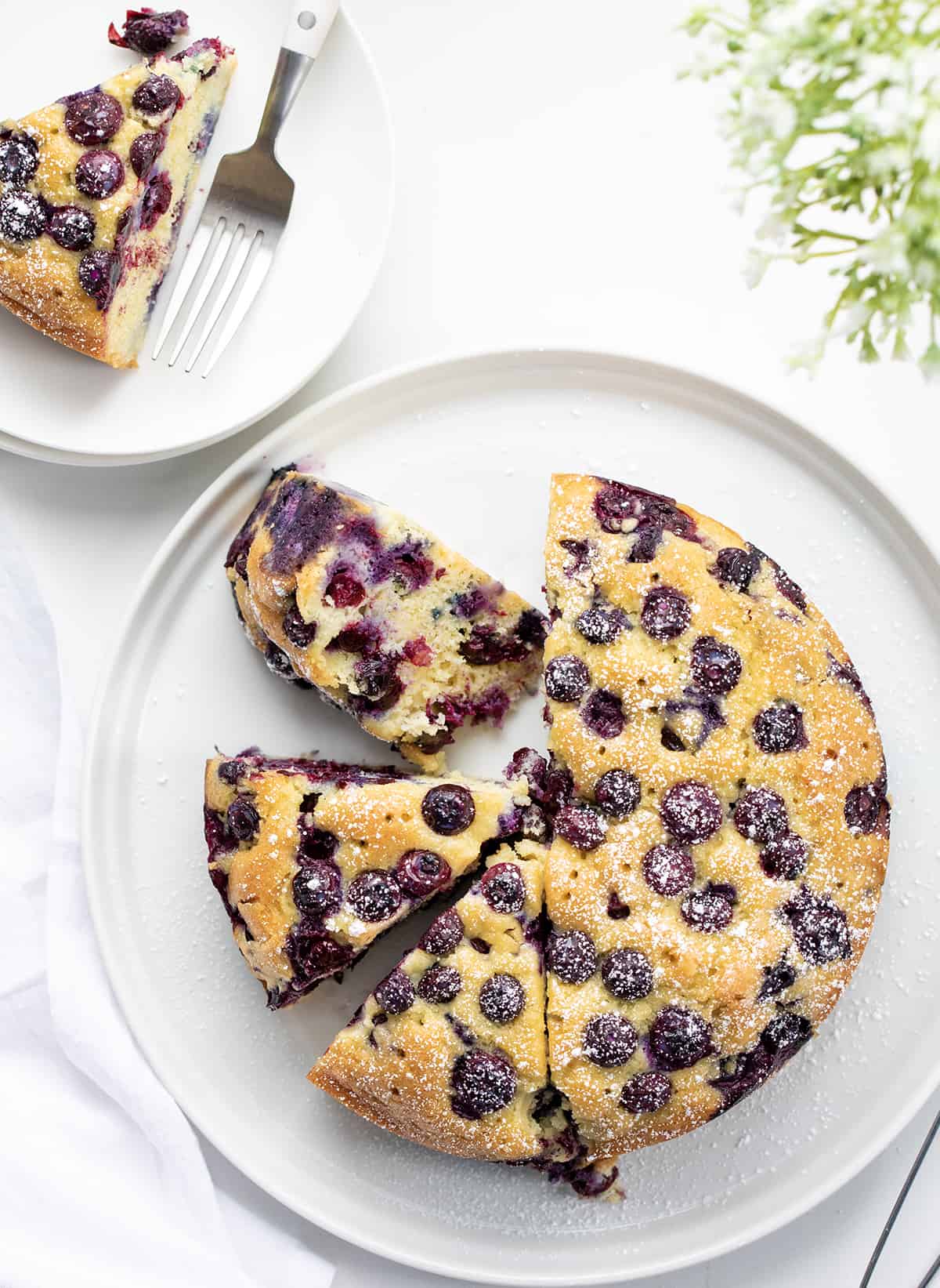 Blueberry Breakfast Cake is loaded with fresh blueberries, baked, and poked with holes to be saturated with a warm sugary milk mixture for a super moist and rich cake. Then, it's dusted with confectioners' sugar. This type of cake, known as a poke cake, is the perfect way to add flavor and extra moisture to each bite. And, don't save this cake just for breakfast; it is also a delicious snack or dessert. Whole Blueberry Breakfast Cake With Some Pieces Cut and One Piece Removed.