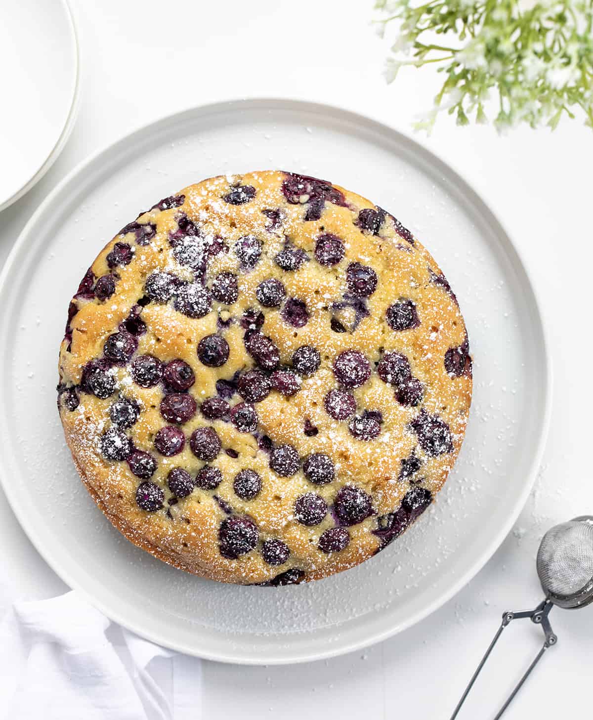 Blueberry Breakfast Cake is loaded with fresh blueberries, baked, and poked with holes to be saturated with a warm sugary milk mixture for a super moist and rich cake. Then, it's dusted with confectioners' sugar. This type of cake, known as a poke cake, is the perfect way to add flavor and extra moisture to each bite. And, don't save this cake just for breakfast; it is also a delicious snack or dessert. Whole Blueberry Breakfast Cake on a White Counter with Flowers.