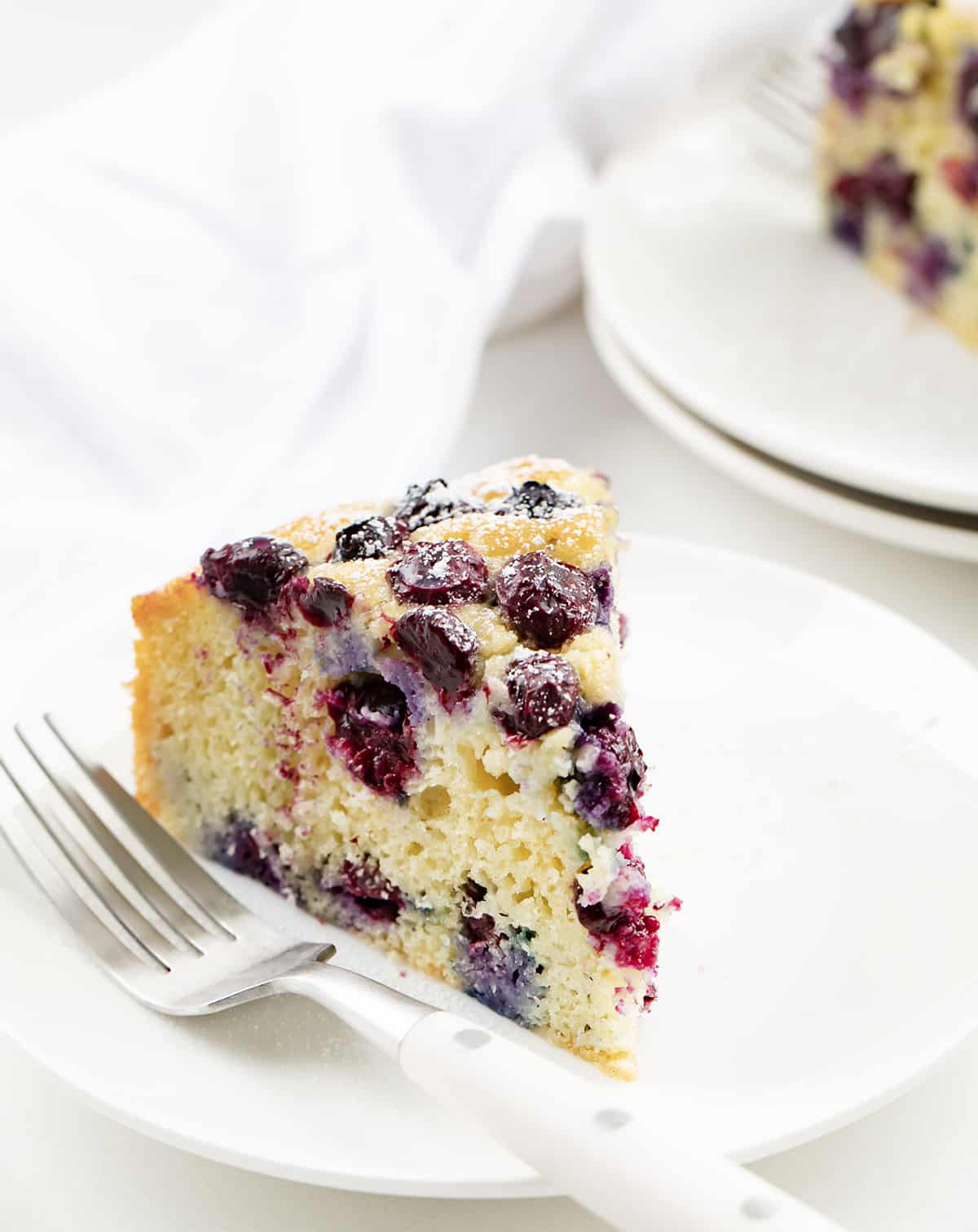 Blueberry Breakfast Cake is loaded with fresh blueberries, baked, and poked with holes to be saturated with a warm sugary milk mixture for a super moist and rich cake. Then, it's dusted with confectioners' sugar. This type of cake, known as a poke cake, is the perfect way to add flavor and extra moisture to each bite. And, don't save this cake just for breakfast; it is also a delicious snack or dessert. Slices of Blueberry Breakfast Cake on White Plates.