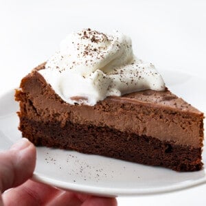 Hand Holding White Plate with Brownie Bottom Chocolate Cheesecake on it.