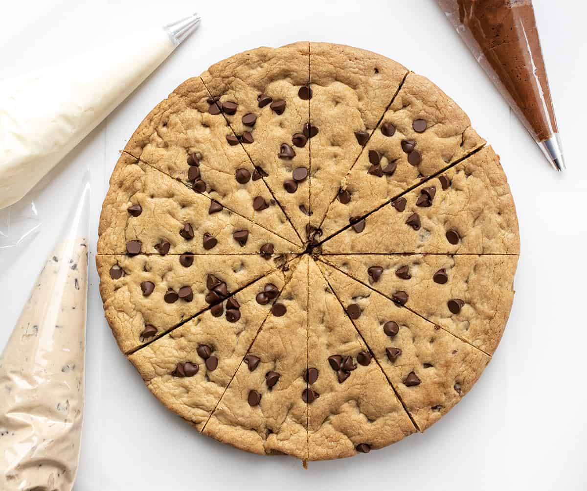 Baked Cookie Cake Cut into 12 Pieces with Vanilla, Chocolate, and COokie Dough Frosting in Piping Bags.