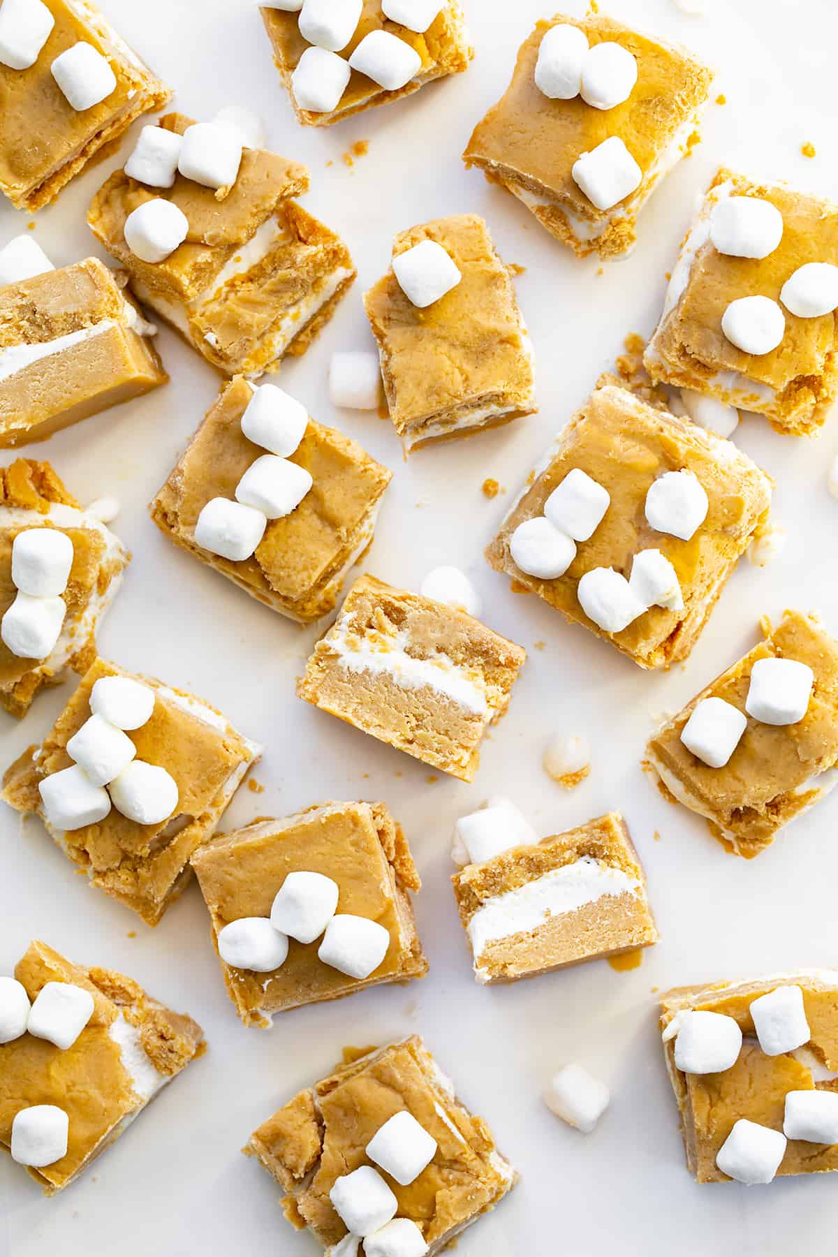 This fudge was inspired by a fluffernutter sandwich, which is a sandwich made with peanut butter and marshmallow fluff. Fluffernutter Fudge is two layers of melted peanut butter and white chocolate with a layer of marshmallow fluff in between. If you love peanut butter, do not miss out on this recipe! Individual Pieces of Fluffernutter Fudge (Peanut Butter Fudge and Marshmallow Fluff) Cut Up and On a White Surface with Some Pieces on Their Side Showing the Layers.