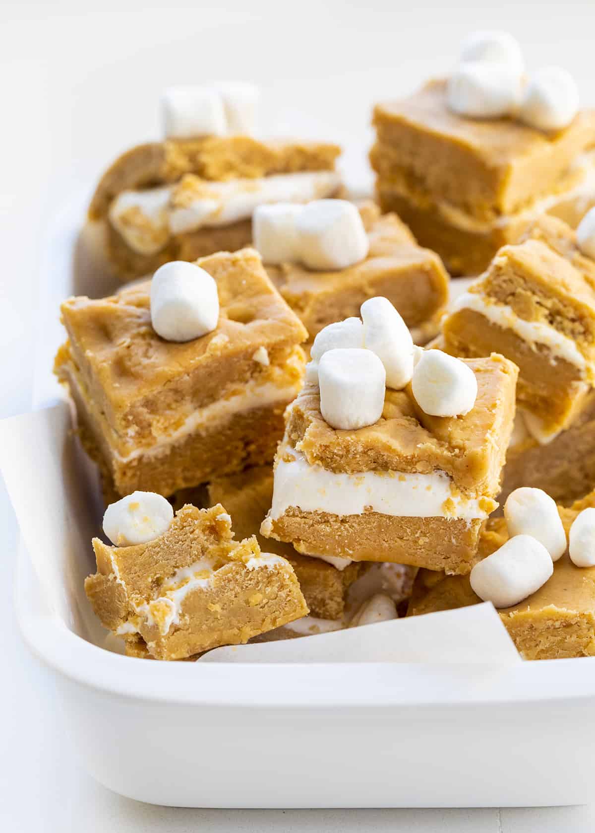 This fudge was inspired by a fluffernutter sandwich, which is a sandwich made with peanut butter and marshmallow fluff. Fluffernutter Fudge is two layers of melted peanut butter and white chocolate with a layer of marshmallow fluff in between. If you love peanut butter, do not miss out on this recipe! Individual Pieces of Peanut Butter and Marshmallow Fluff Fudge also known as Fluffernutter Fudge in a White Baking Dish and Mini Marshmallows on Top.