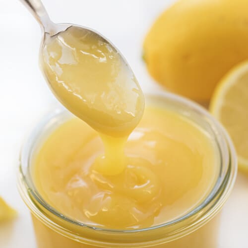 Spooning out some fresh Lemon Curd from a Jar surrounded by Lemons.