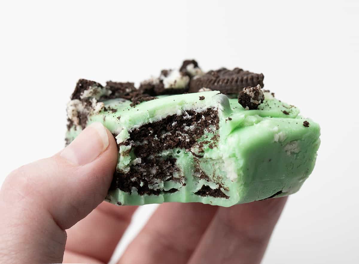 Creamy, no-fuss Mint Chocolate Oreo Fudge needs to be the next recipe on your dessert line-up. This delicious mint and chocolate cookie treat will have your guests coming back for seconds and thirds. Hand Holding a Piece of Mint Oreo Fudge with a Bite Taken Out.