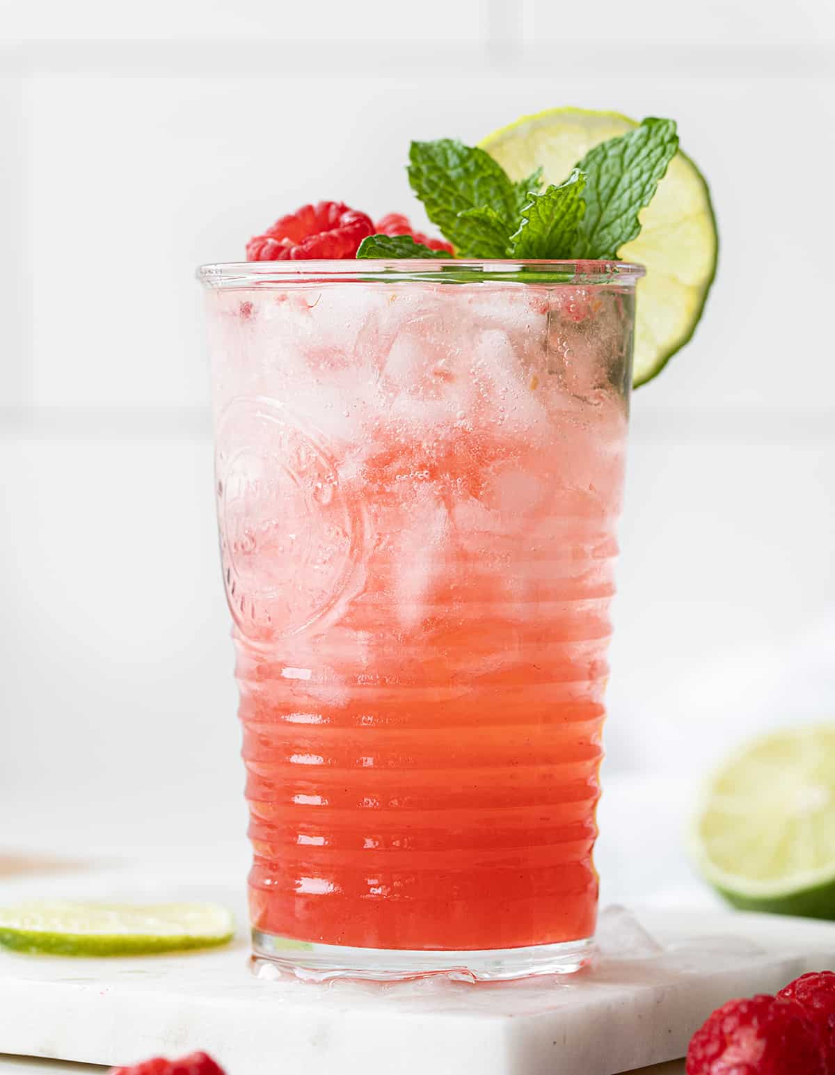 One Glass of Raspberry Lime Mojito on a Mini Marble Cutting Board with Limes and Raspberries Around it.