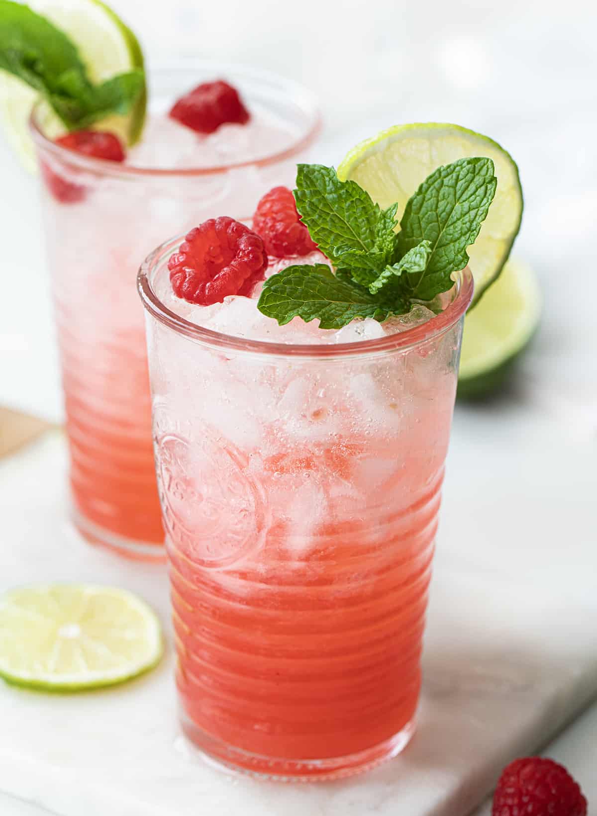 Two Glasses of Raspberry Lime Mojito on a Mini Marble Cutting Board with Limes and Raspberries Around it.