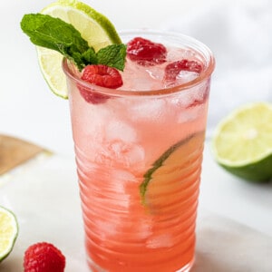 One Glass of Raspberry Lime Mojito on a Mini Marble Cutting Board with Limes and Raspberries Around it.