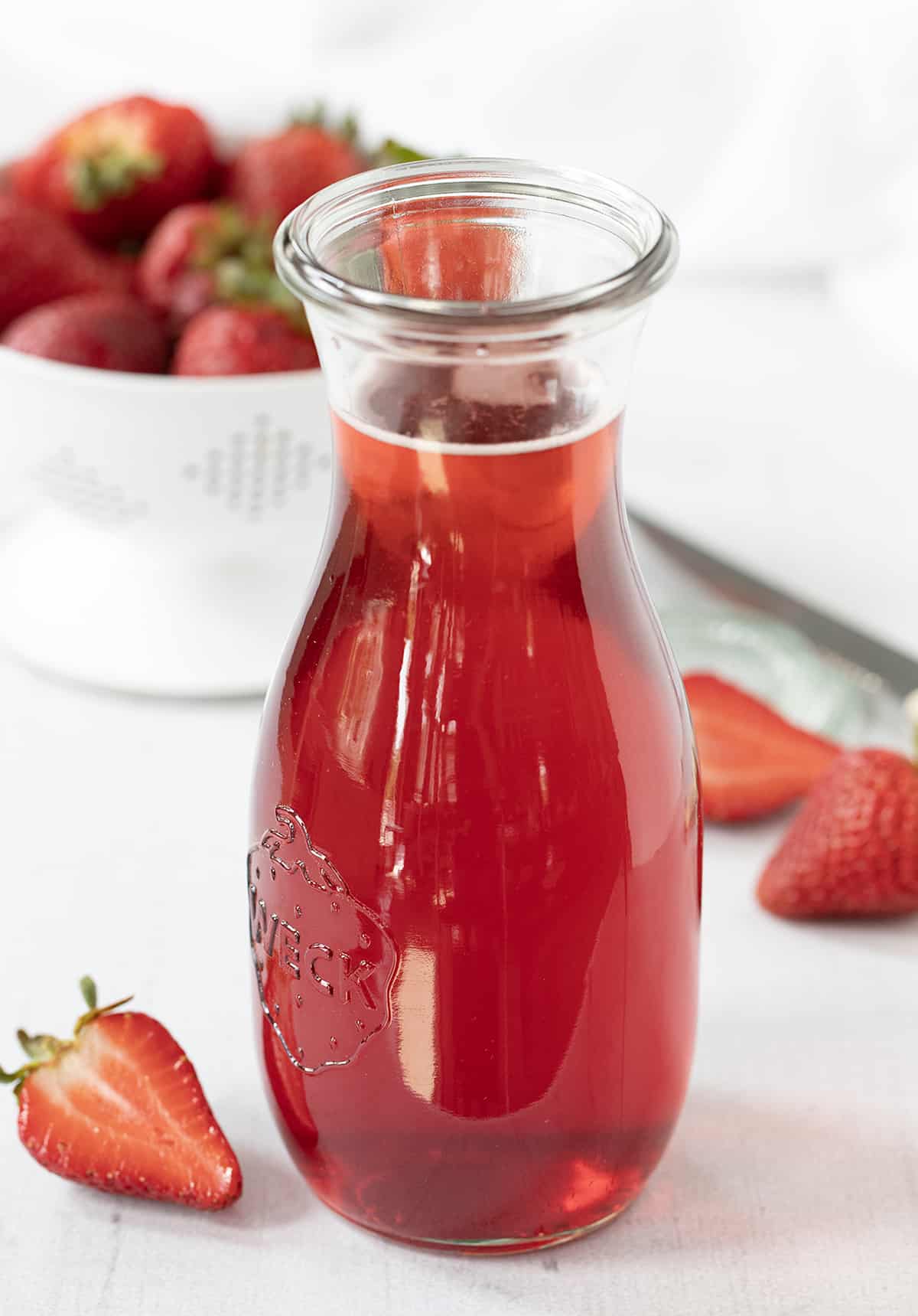 Strawberry Simple Syrup is a homemade sweetener made with just three ingredients, sugar, water, and fresh strawberries. It can add a sweet and fruity flavor to a variety of recipes from beverages to desserts to salads. If you can boil water, you can make simple syrup! Bottle of Strawberry Simple Syrup Surrounded by Strawberries.