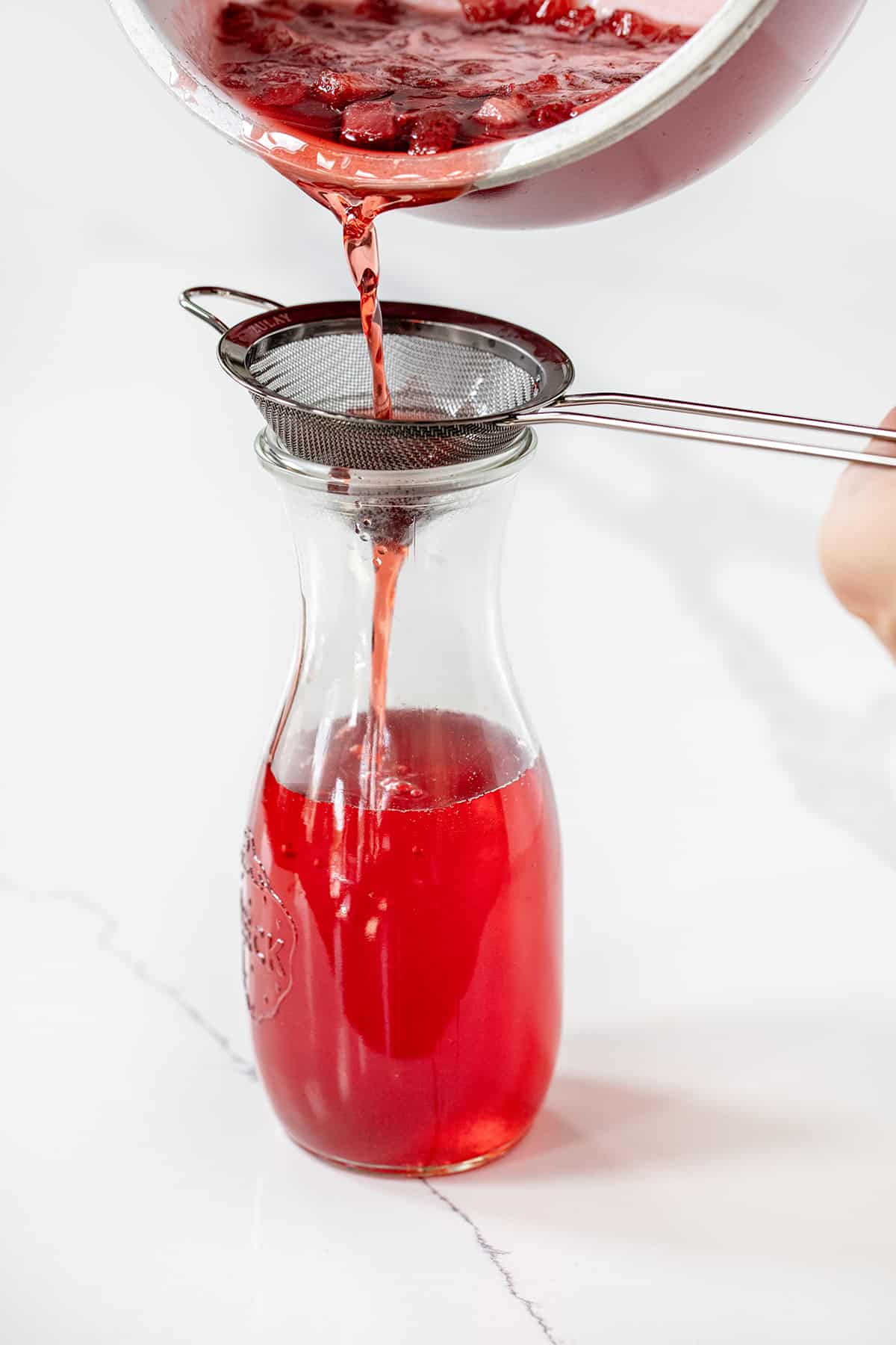 Strawberry Simple Syrup is a homemade sweetener made with just three ingredients, sugar, water, and fresh strawberries. It can add a sweet and fruity flavor to a variety of recipes from beverages to desserts to salads. If you can boil water, you can make simple syrup! Straining Strawberry Mixture into Bottle to Make Strawberry Simple Syrup.