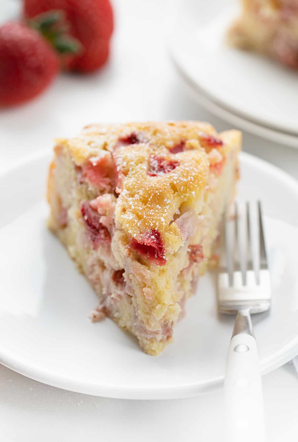 Piece of Fresh Strawberry Cake on a White Plate with Strawberries in the Background.