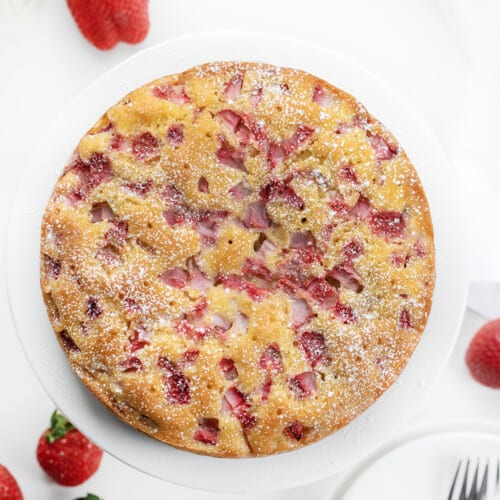 Whole Fresh Strawberry Cake on a White Plate with Strawberries in the Background.