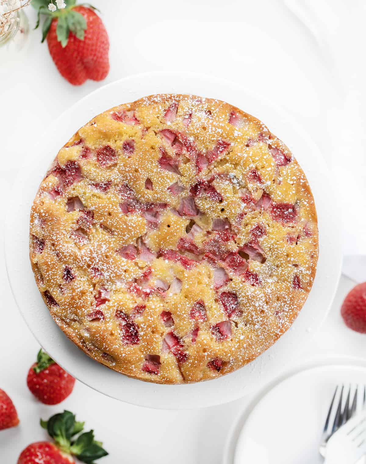 Whole Fresh Strawberry Cake on a White Plate with Strawberries in the Background.